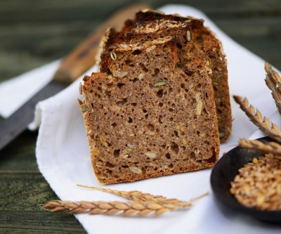 Ein angeschnittenes Vollkornbrot liegt auf einem Tuch, man sieht die volle Bandbreite an Körnern und Samen, davor steht eine Schüssel mit Getreidekörnern und daneben liegt eine Ähre vom Feld. Im Hintergrund sieht man noch leicht das Messer.
