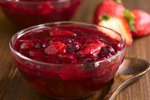 Beerengrütze in einer Glasschüssel auf einem Holztisch. Daneben ein Holzlöffel und geschnittene Erdbeeren